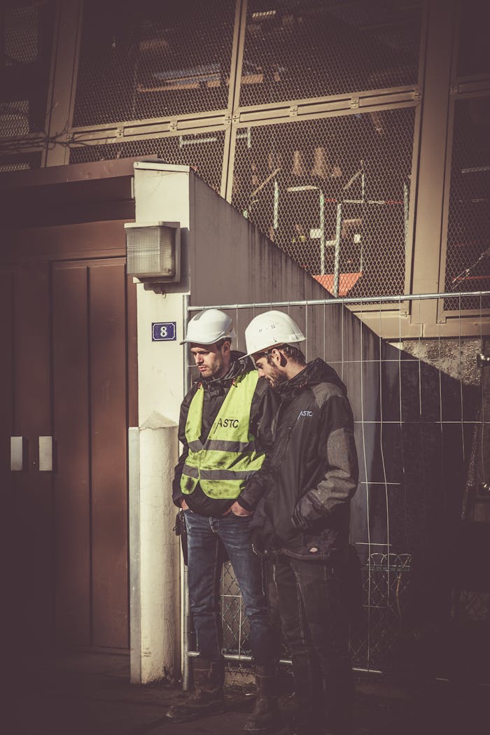 contact-img Two construction workers in safety gear standing outdoors at a building site, focused on work.